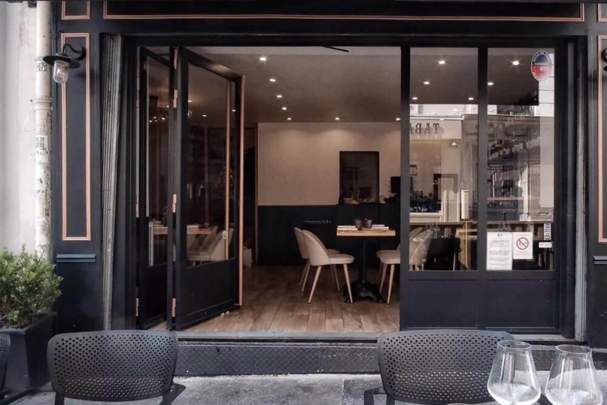 Restaurant exterior with black storefront, open doors showing wooden tables inside, terrace chairs out front