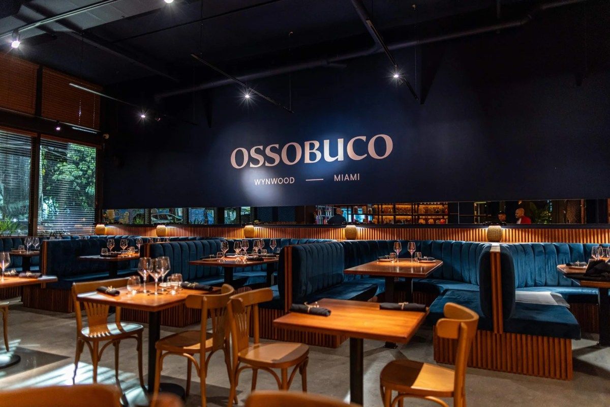 Interior dining room of Ossobuco Wynwood Miami with blue booth seating and wooden tables, Ossobuco sign on wall