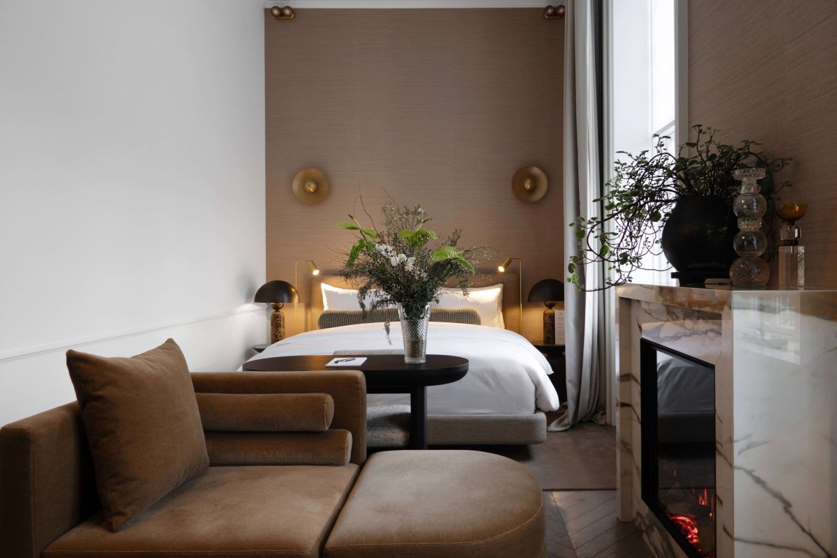 Elegant hotel suite with bed, brown velvet armchair, marble fireplace, and fresh flowers on a side table