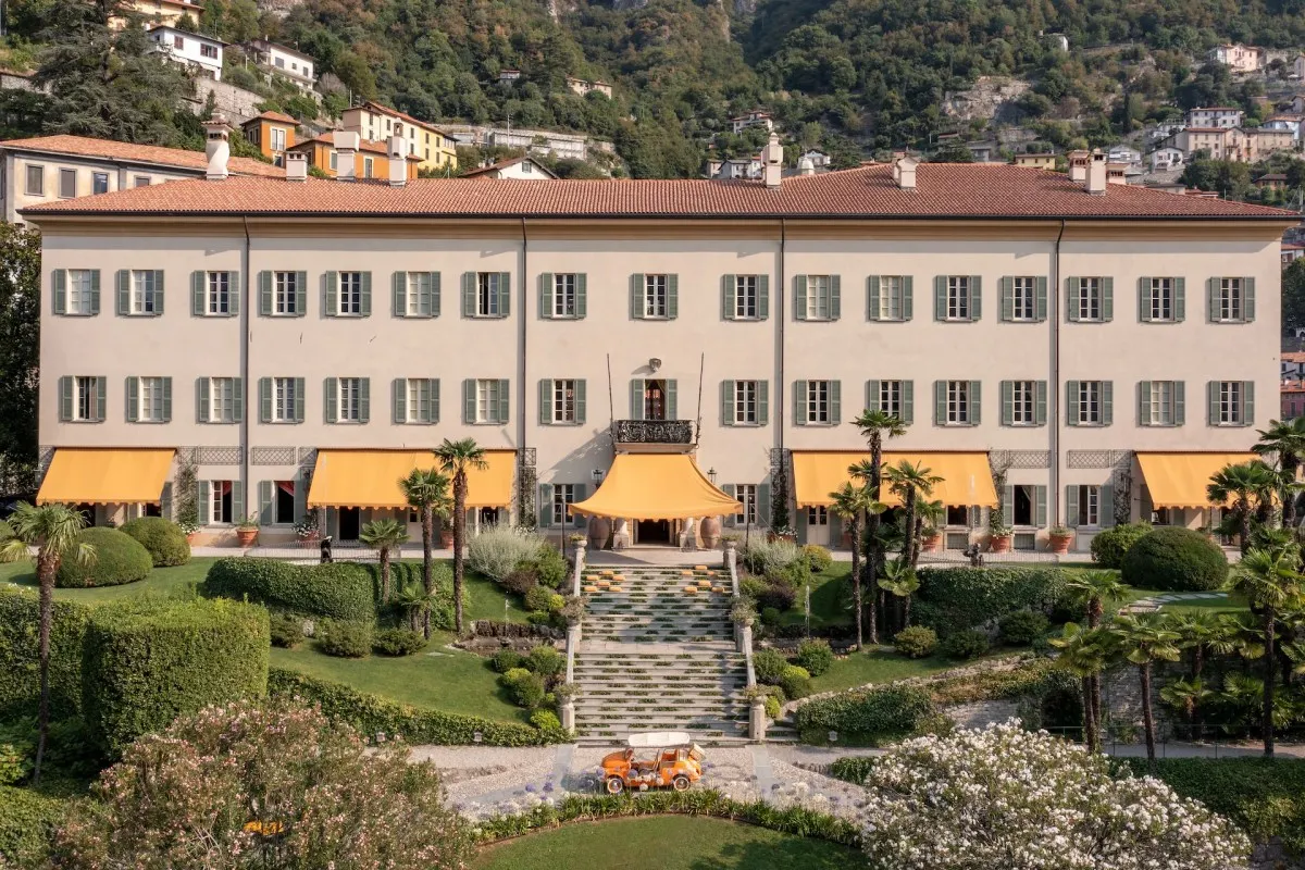 The cream façade of Villa Passalacqua with green shutters and orange awnings above terraced gardens and a central stone staircase in Moltrasio
