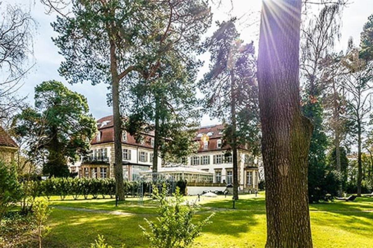 White mansion-style hotel building with red tiled roof seen through tall trees in a sunny park setting