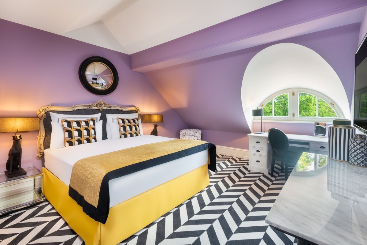 Whimsical purple guest suite with yellow and black striped bedspread, chevron floor pattern, and an arched window overlooking greenery.