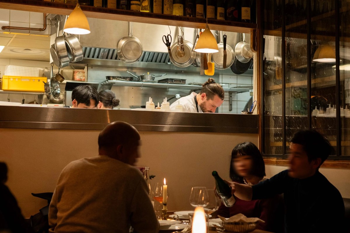 Restaurant dining room interior with diners at a candlelit table and an open kitchen with chefs working in the background.
