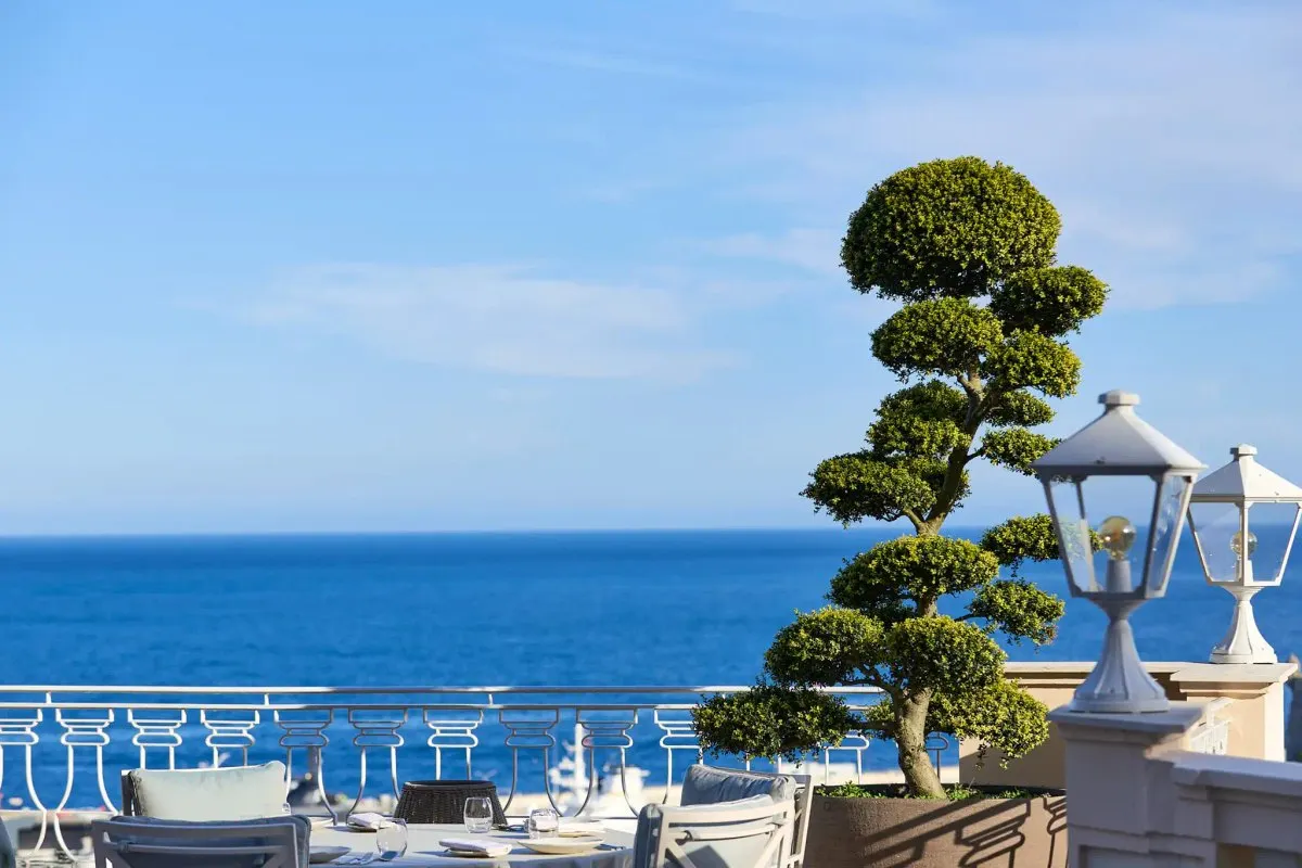 The terrace of Pavyllon Monte-Carlo at Hôtel Hermitage, with laid tables, a clipped topiary tree and lanterns above the blue Mediterranean