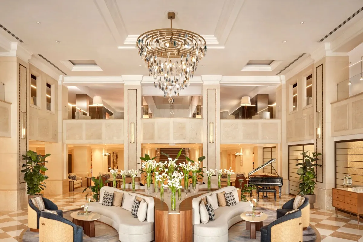 Grand two-story hotel lobby with marble checkered floors, ornate chandelier, white curved sofas, and piano