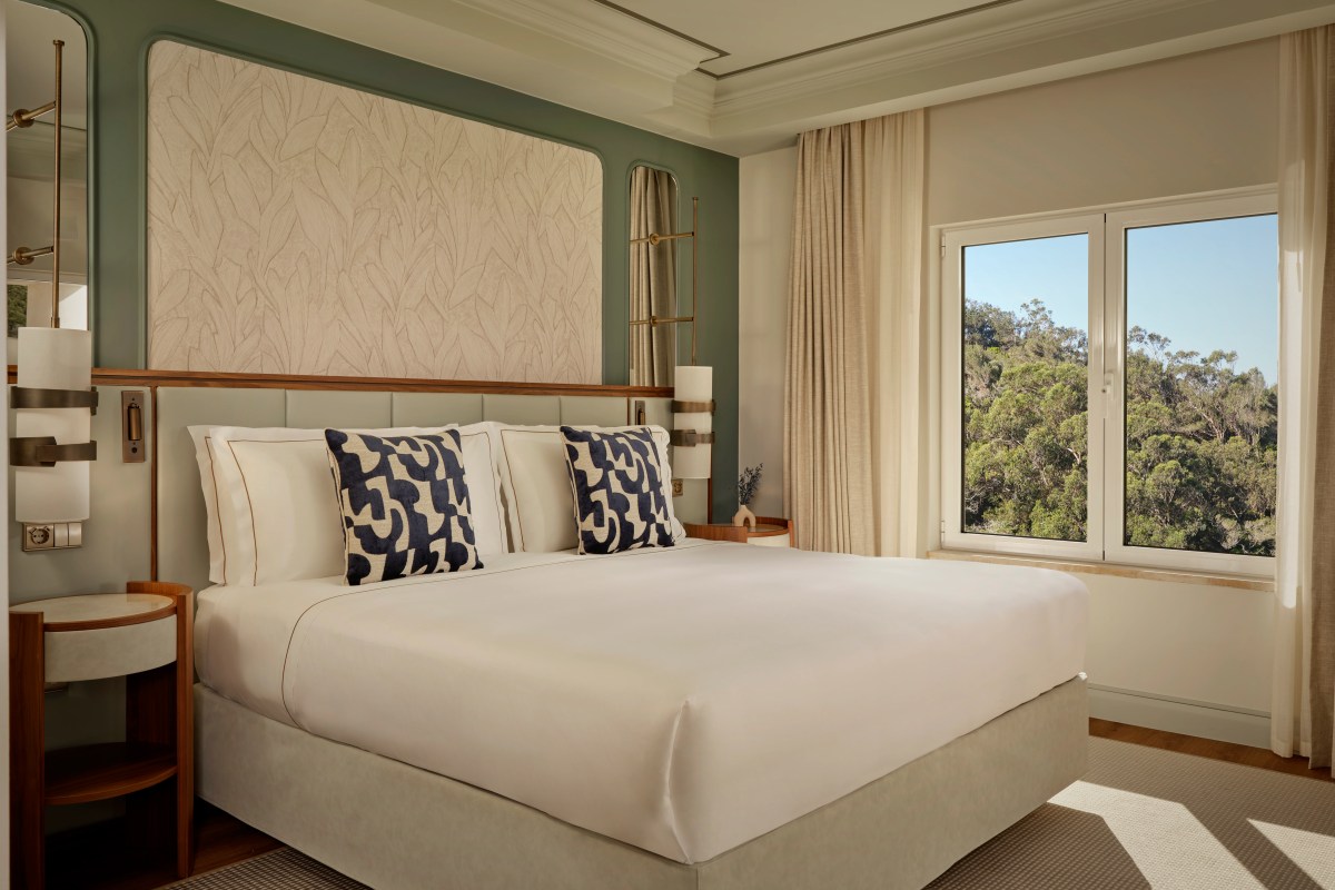 Hotel bedroom with king bed, sage green headboard, patterned pillows, and large window overlooking trees