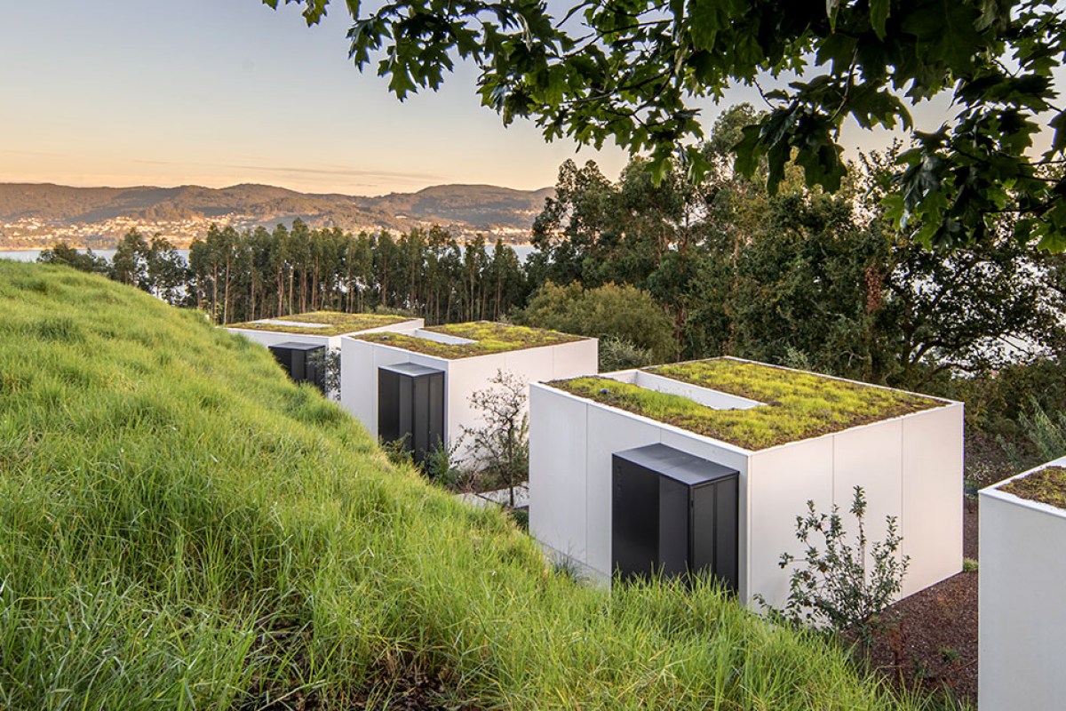 Row of modern white hotel pavilions with green roofs on a grassy hillside, overlooking trees and a bay at sunset
