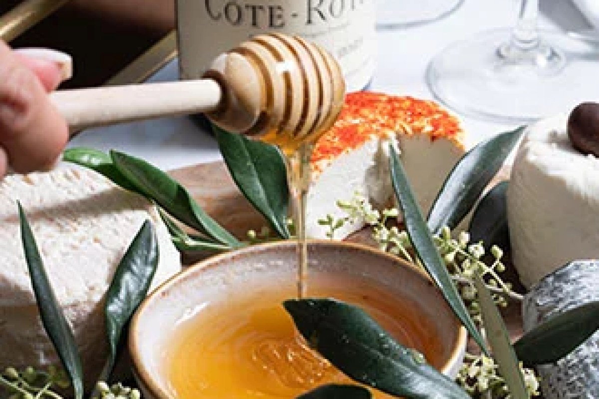 Close-up of cheese board with honey being drizzled from a dipper, olive leaves, and a wine bottle