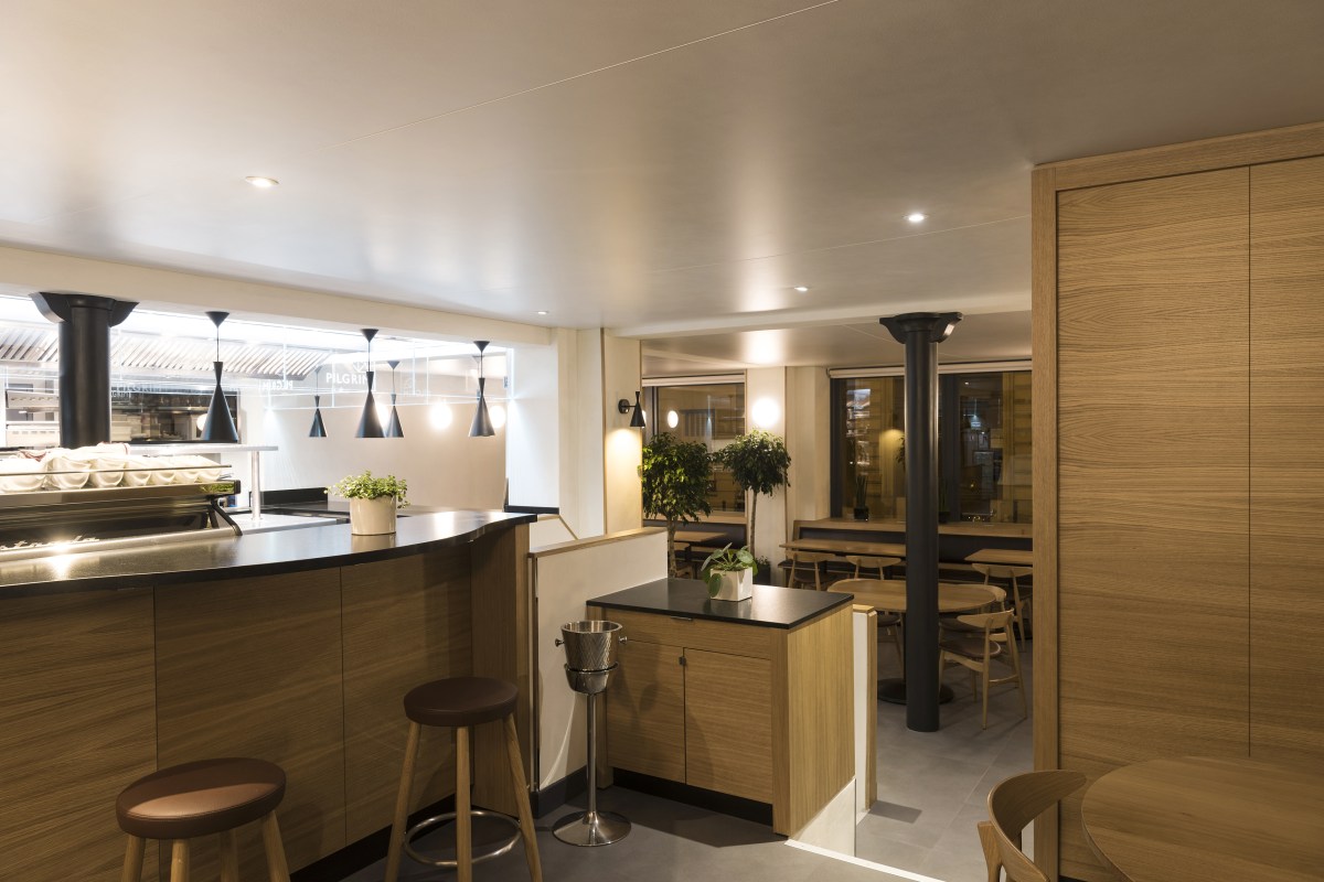 Wood-paneled bar and dining area with stools, plants, and tables, warmly lit interior view.
