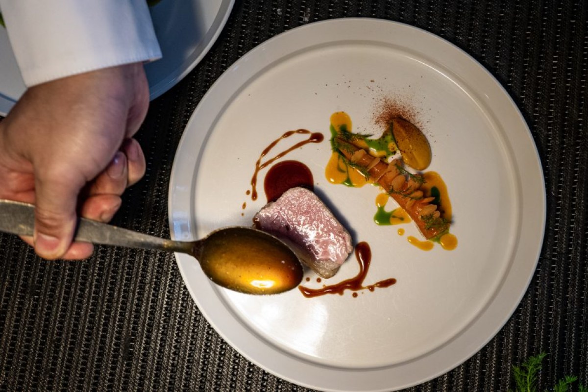 Overhead shot of a chef's hand spooning sauce onto a plated dish with pork, carrot, and herb garnish.