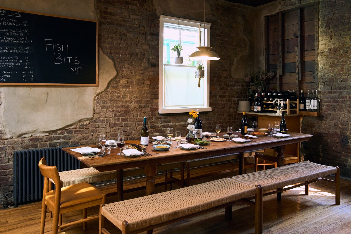 Rustic dining room with exposed brick walls, a long wooden table set with wine bottles and dishes, chalkboard menu, and a wine rack