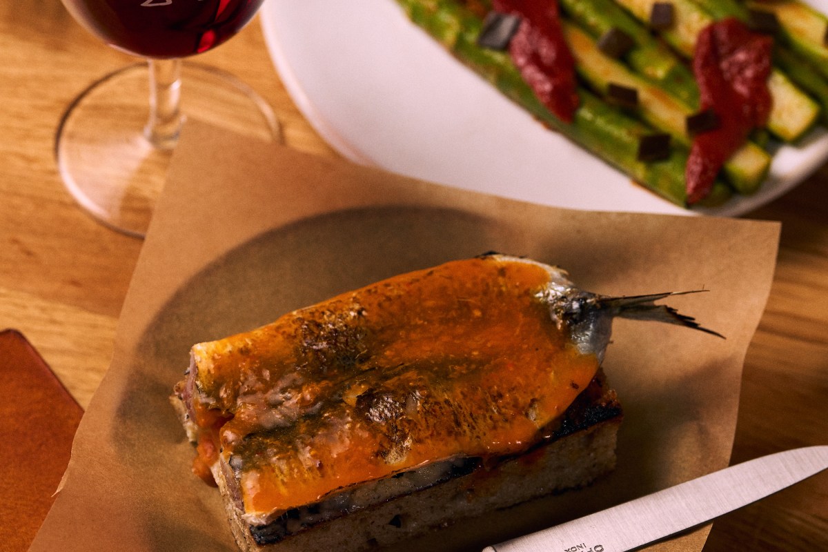 Grilled sardine toast with orange sauce on brown paper, glass of red wine and a plate of charred zucchini in the background