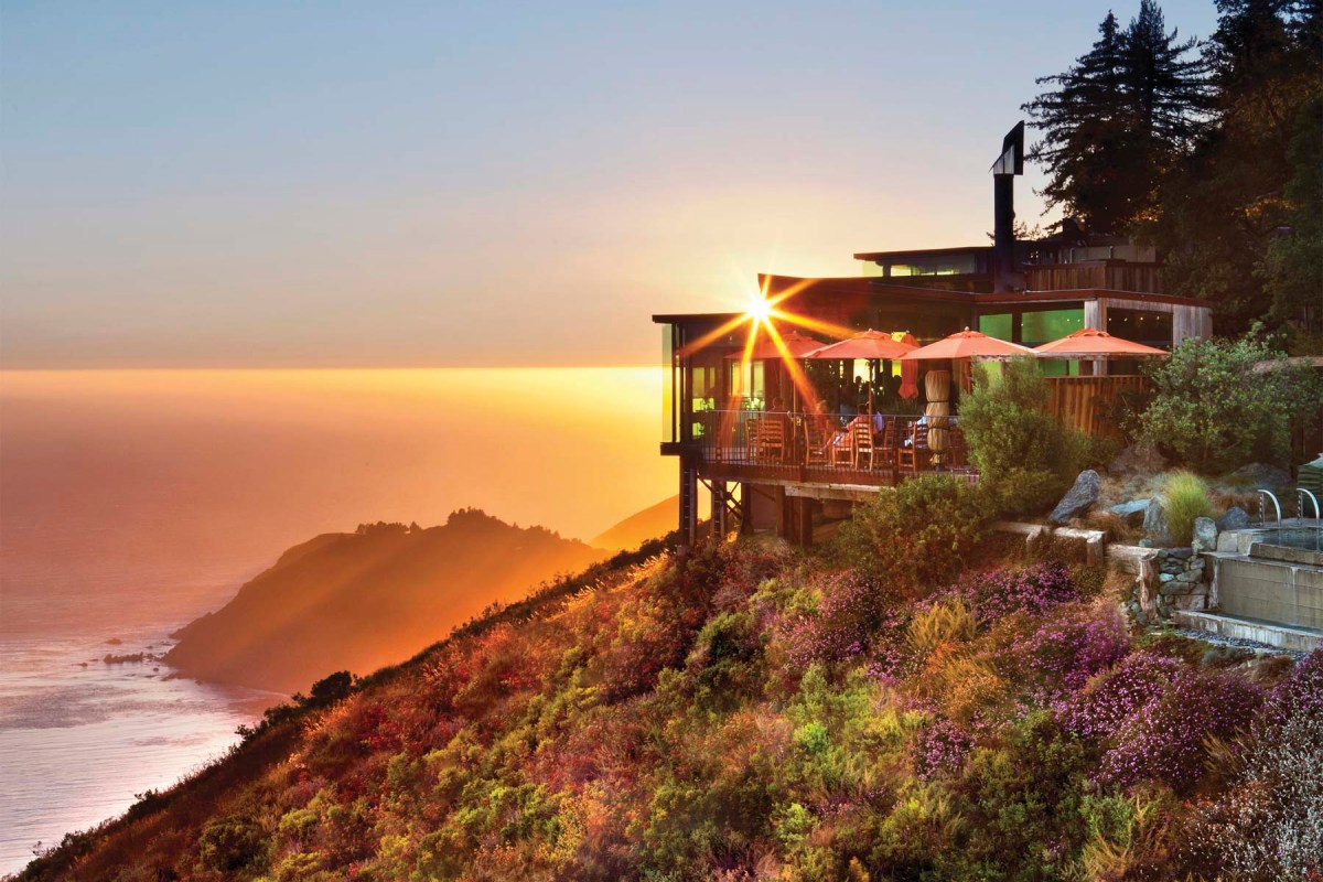 Post Ranch Inn building perched on a cliff at sunset, ocean and coastal hills in the background, sunburst over the roofline