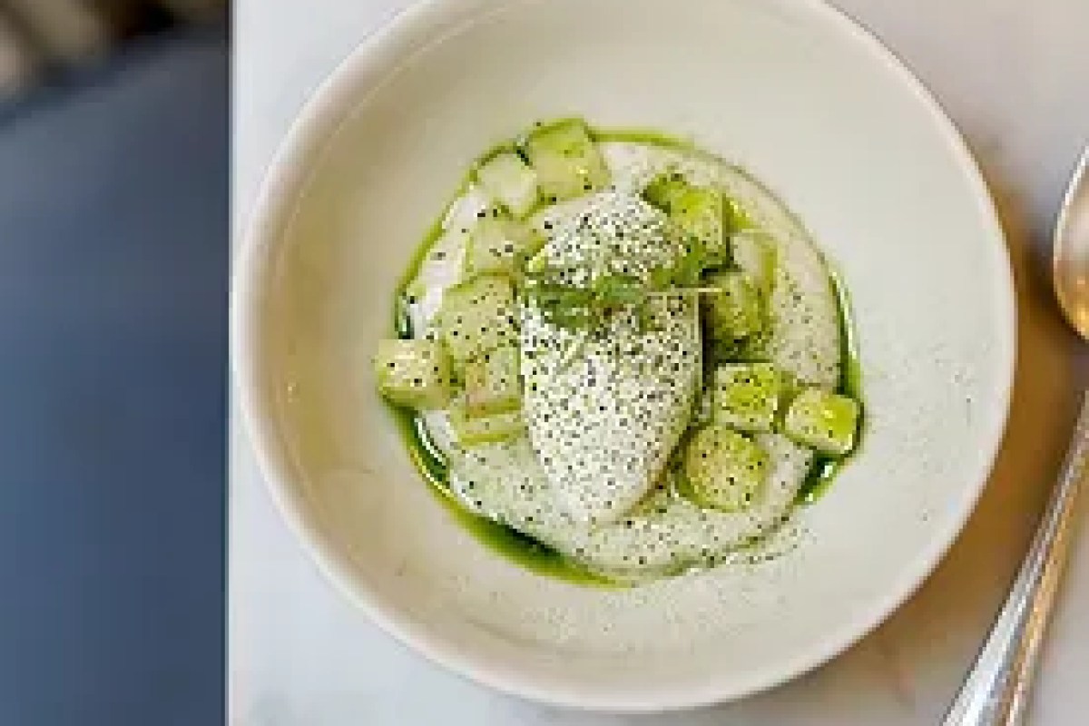 Minimalist white dish with cubed green fruit, a poached element, and herb garnish, served with a spoon on a marble table
