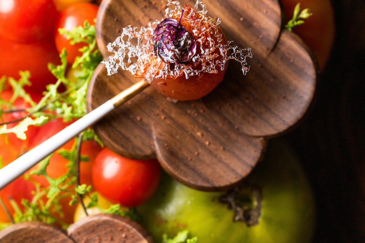 Close-up of a gourmet dish with tomatoes, sugar lace, and purple petal garnish on a wooden flower-shaped board