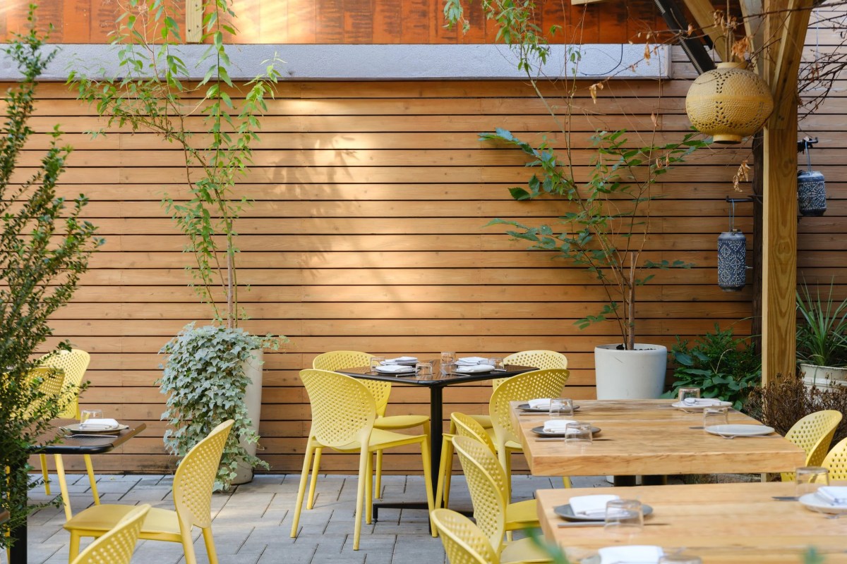 Outdoor dining terrace with yellow chairs, wooden tables, wood-slat wall, and hanging lanterns