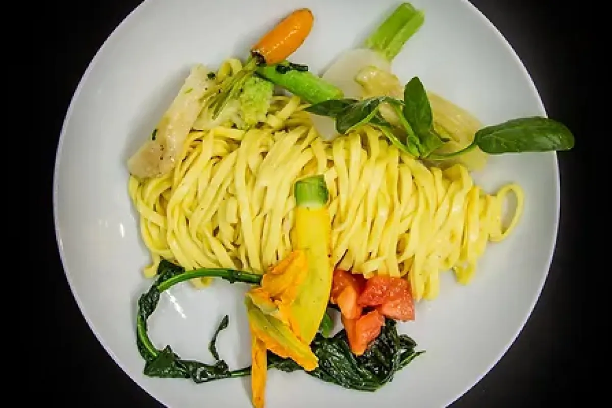 Overhead view of tagliolini pasta with zucchini flowers, vegetables and herbs on a white plate
