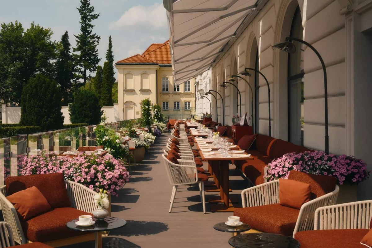 Elegant outdoor terrace with long dining tables, orange cushioned seating, and flowering plants along the hotel's façade