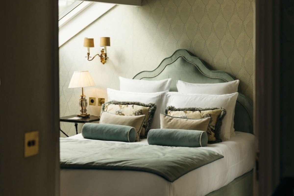 Elegant guest room bed with sage green velvet headboard, bolster pillows and bedside lamp under skylight
