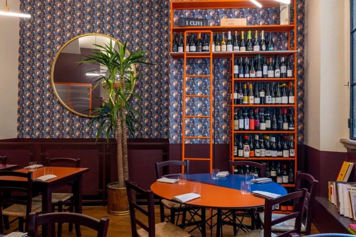 Interior dining room with patterned wallpaper, wine shelf, round mirror and colorful tables set for dining