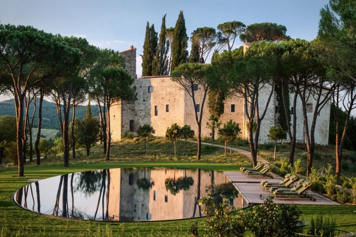 Stone castle hotel building surrounded by pine and cypress trees with reflecting pool and lounge chairs in foreground