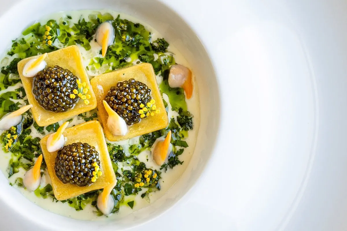 Squares of ravioli topped with caviar on a bed of green herbs in a white bowl, a dish by Julien Roucheteau at Le Restaurant des Rois