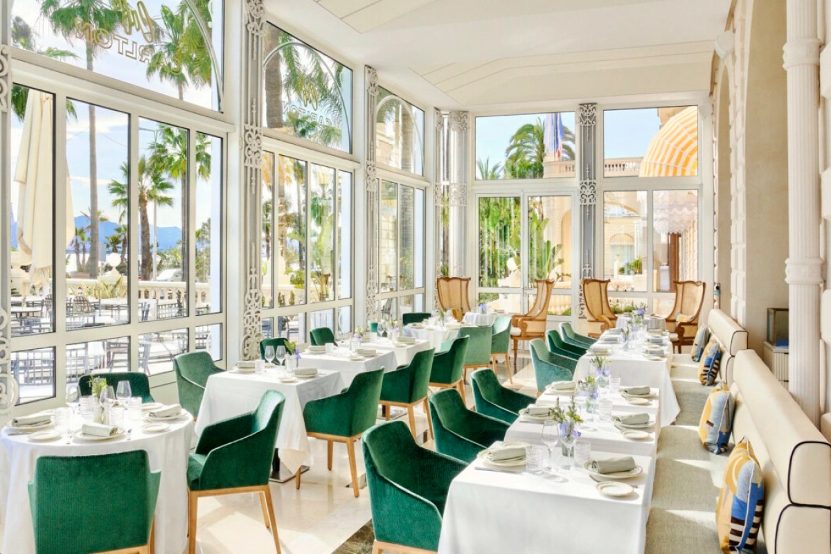Glass-walled dining room with green velvet chairs, white table settings and palm tree views, Riviera restaurant interior