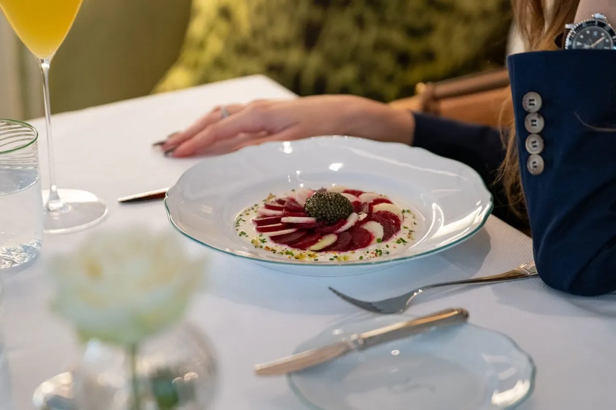 A plate with a thinly sliced starter topped with caviar on a white tablecloth at Robuchon Monaco, beside a glass and a white rose