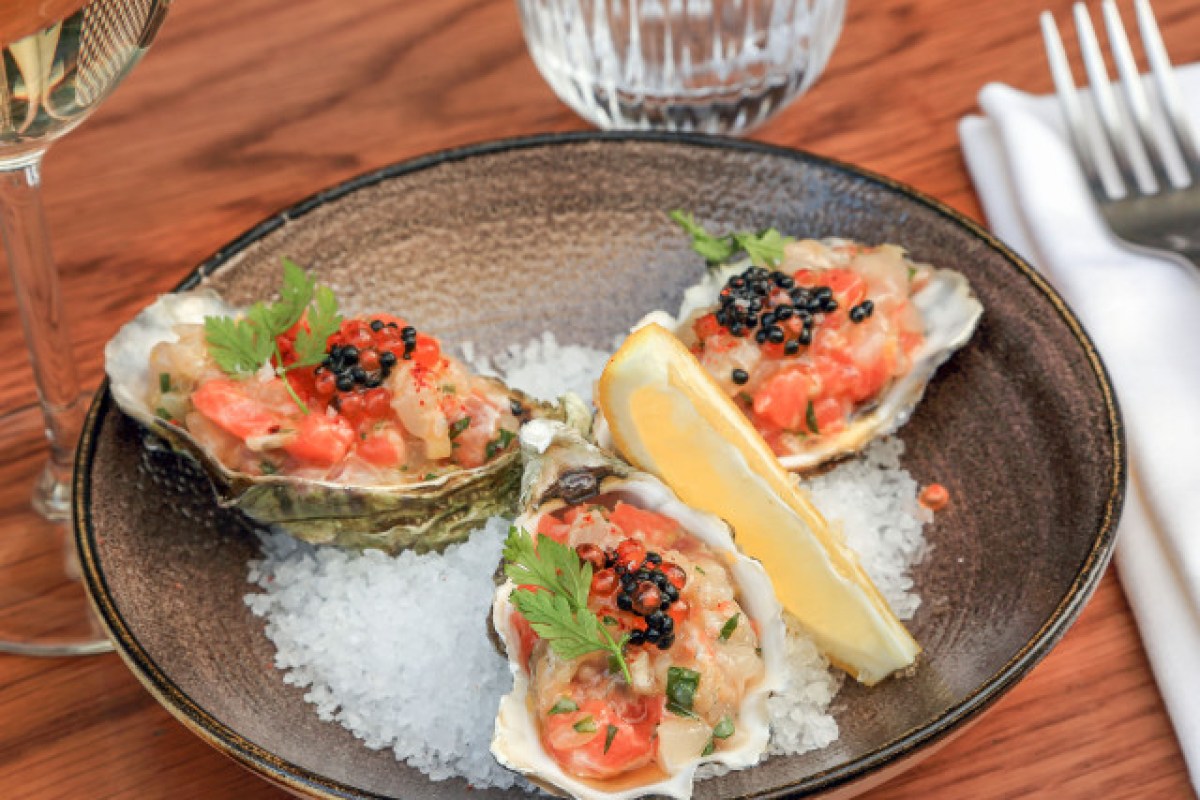 Three oysters topped with diced tomato, caviar and herbs on a plate with lemon, at a restaurant table.