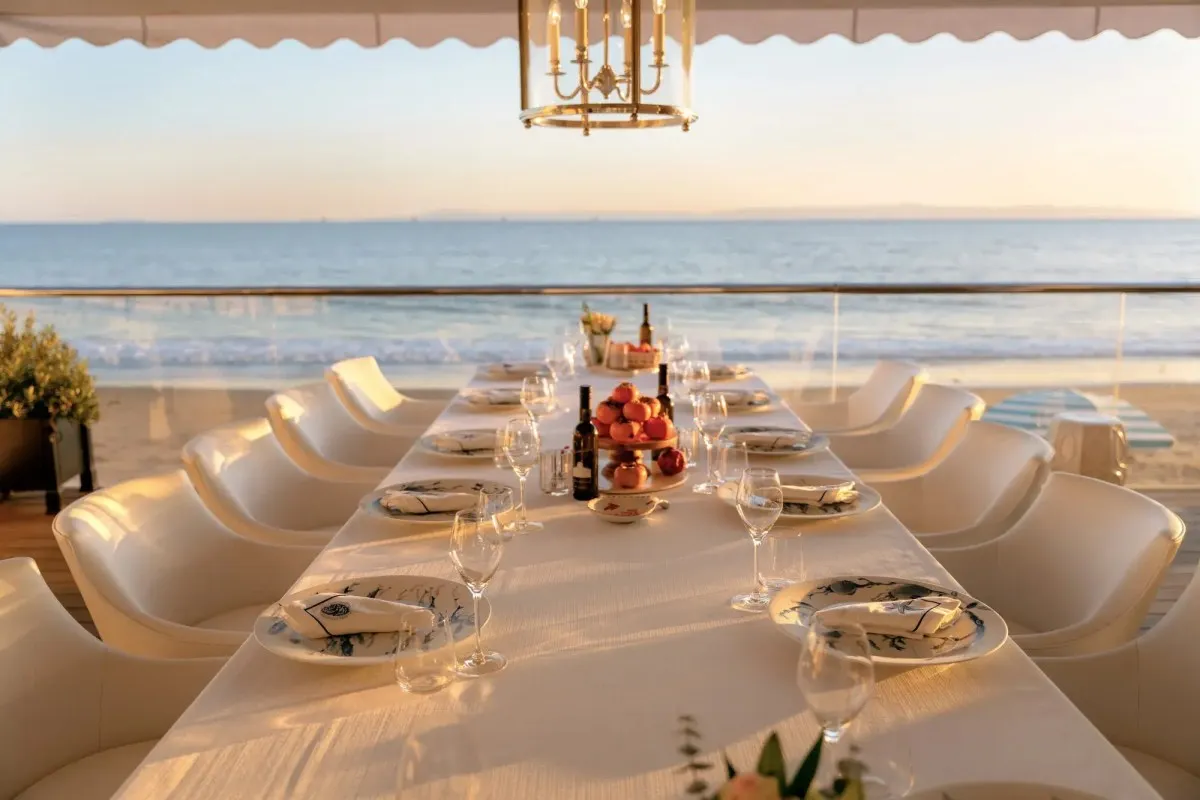 Oceanfront dining table set for a group meal at sunset under an awning, overlooking the beach