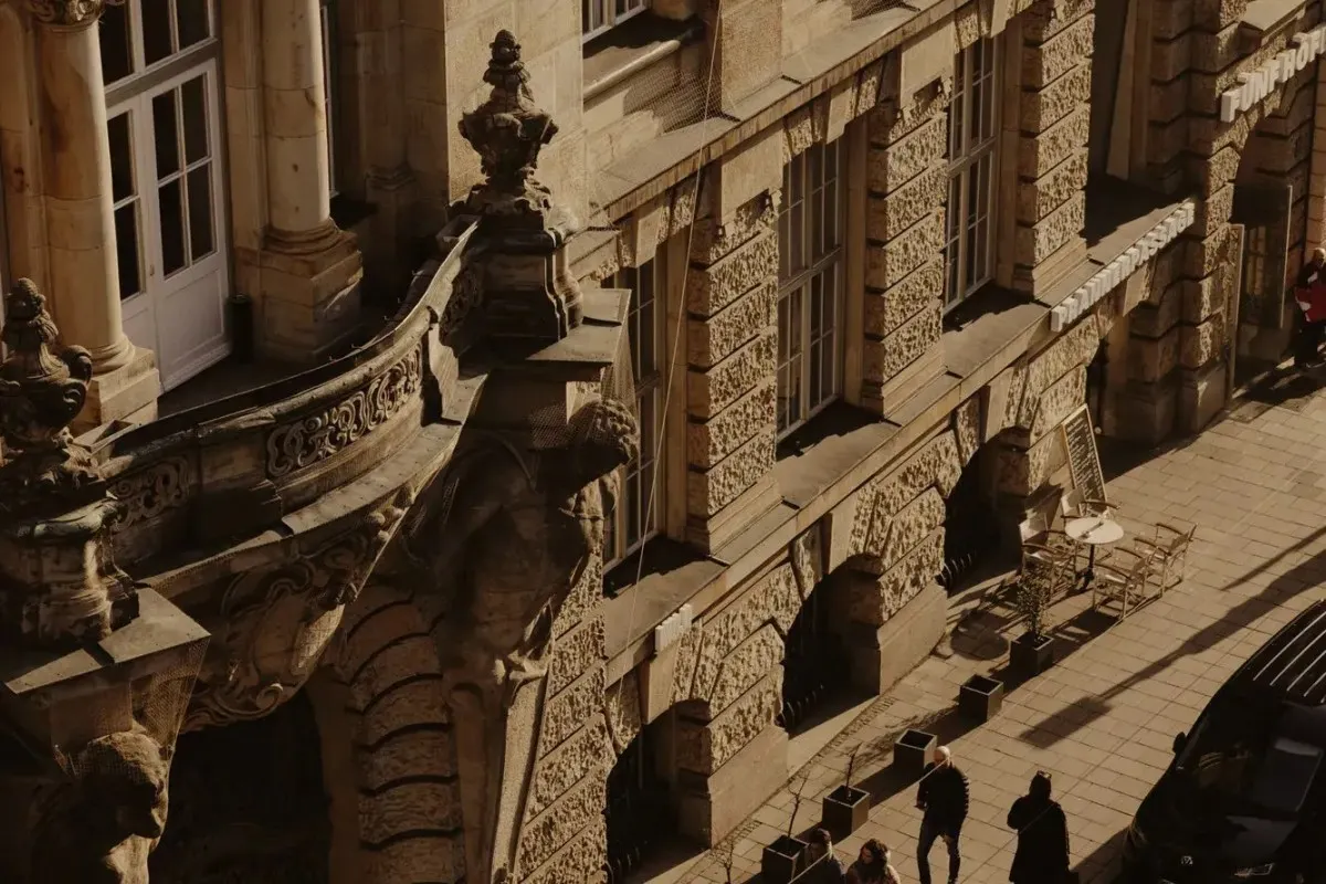 Aerial close-up of the hotel's ornate stone façade with balcony details, windows, and a street-level café terrace below.
