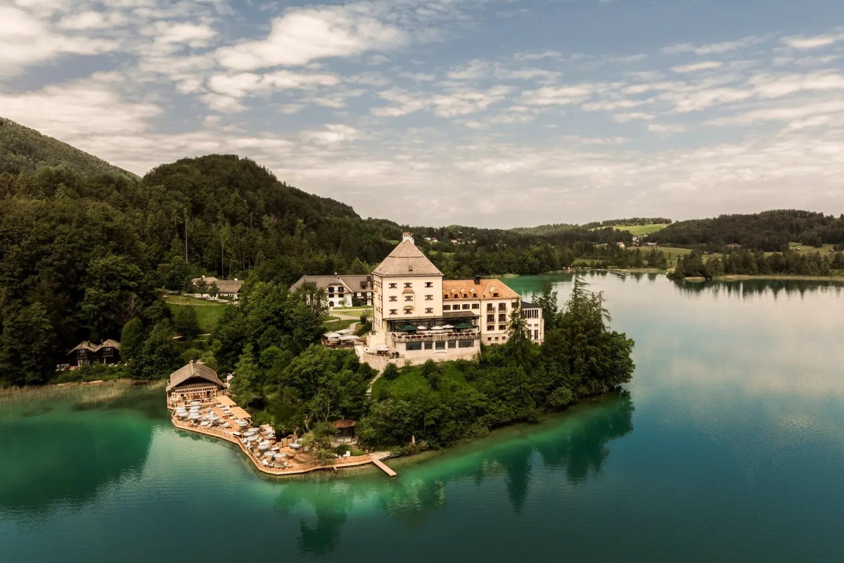 Aerial daytime view of Rosewood Schloss Fuschl castle hotel on a peninsula surrounded by a turquoise lake and forested hills
