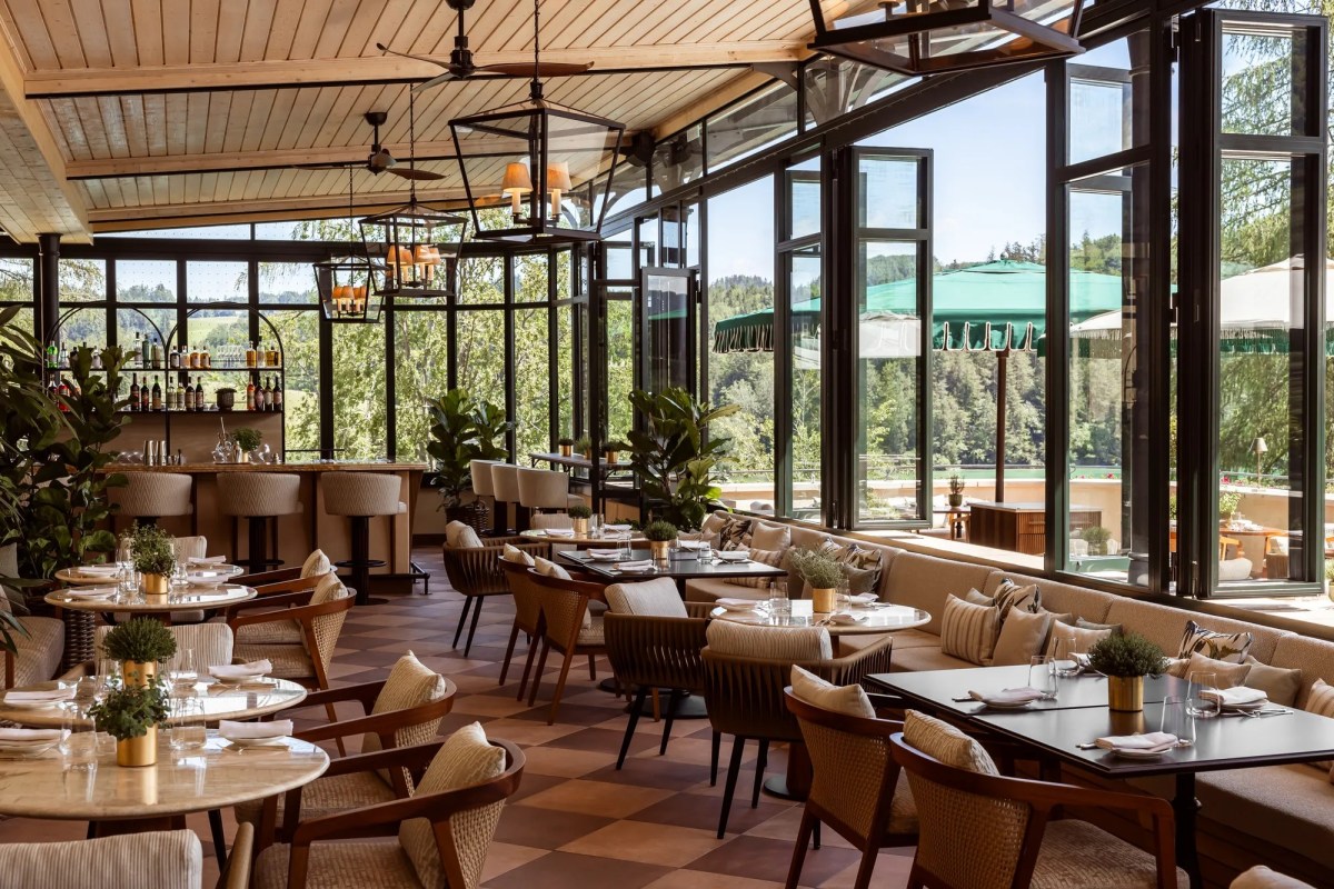 Elegant hotel restaurant terrace with checkered floor, rattan chairs, bar, and large windows overlooking pool and trees