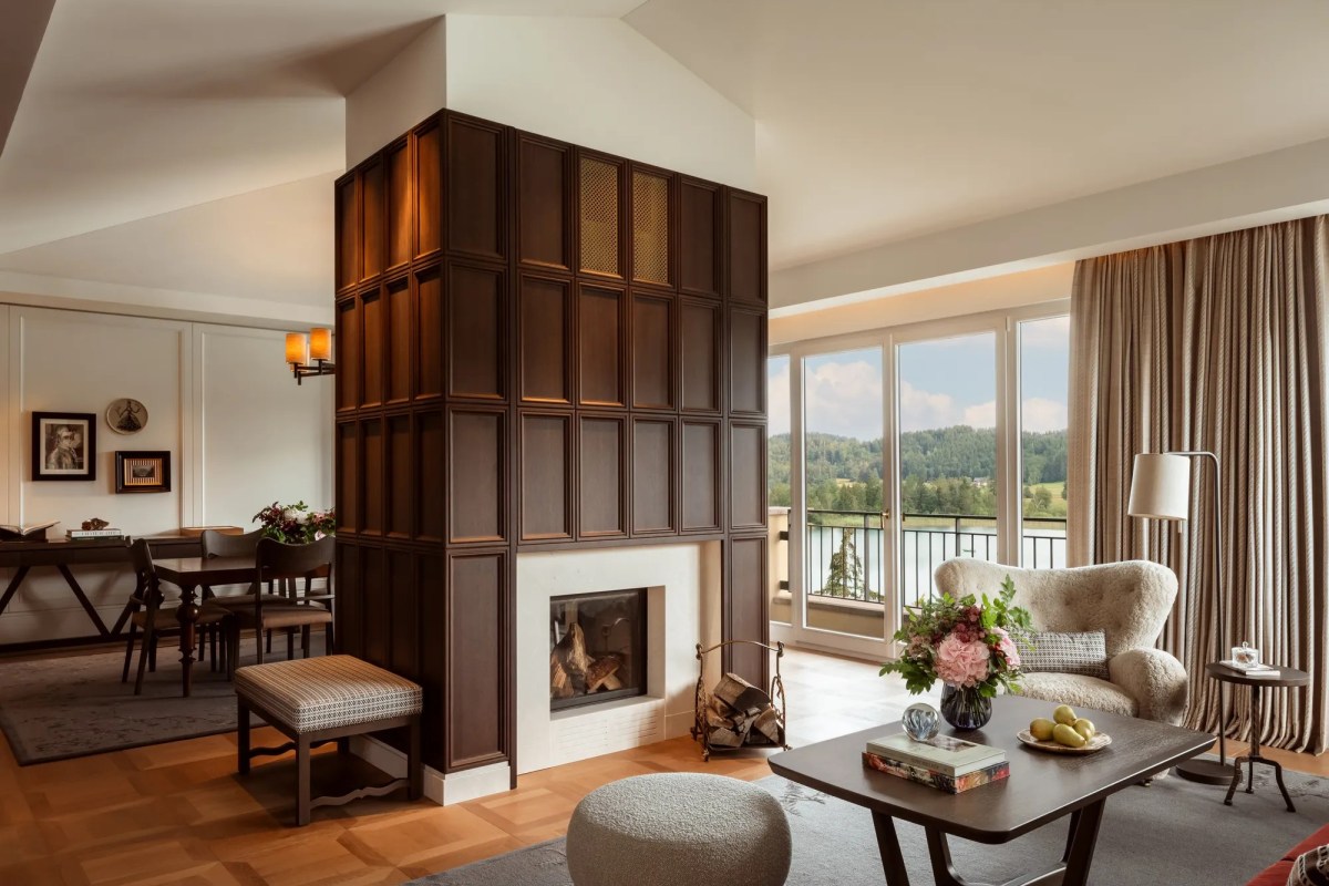 Luxury hotel suite living area with dark wood fireplace surround, armchair, dining table, and lake view through balcony doors