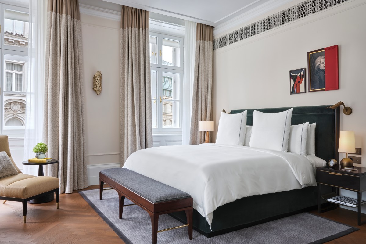 Elegant guest room with dark green upholstered bed, bench, and city view through tall windows