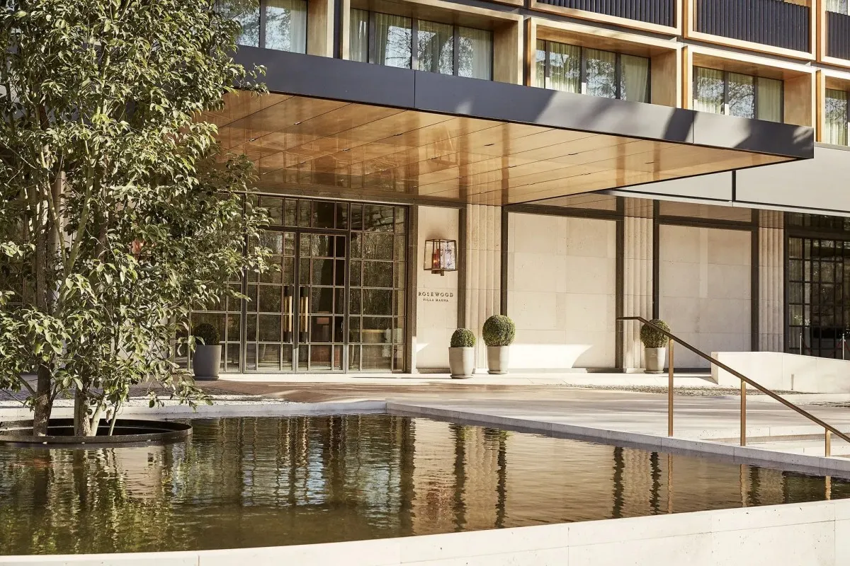 Hotel entrance facade with reflecting pool, tree and golden canopy over glass doors, no people visible.
