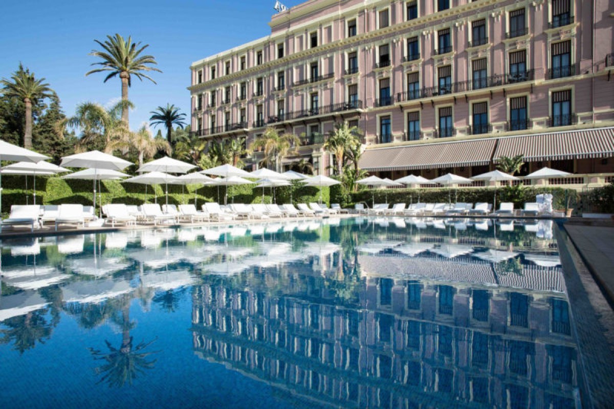 Hotel's pink facade with multiple balconies behind a large pool lined with white loungers and umbrellas
