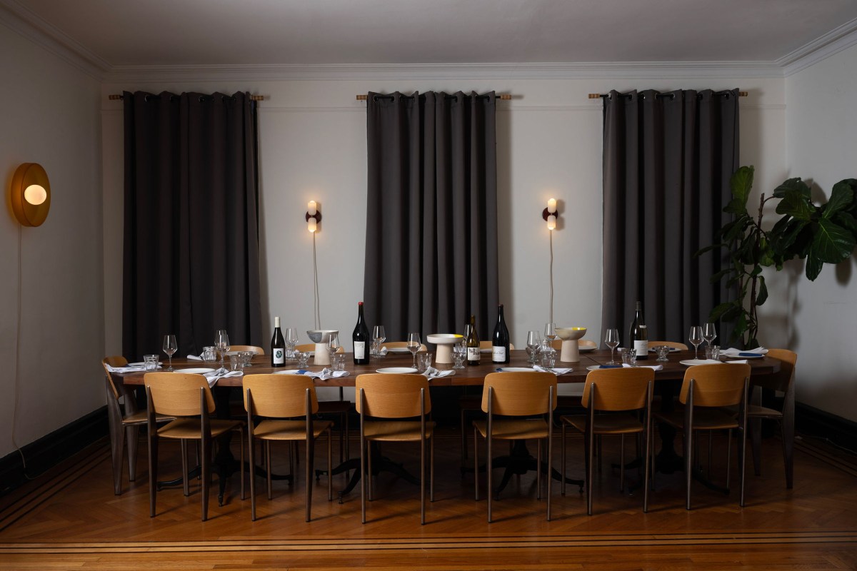 Long wooden dining table set for a group with wine bottles, glasses, and wall sconces against dark curtains.