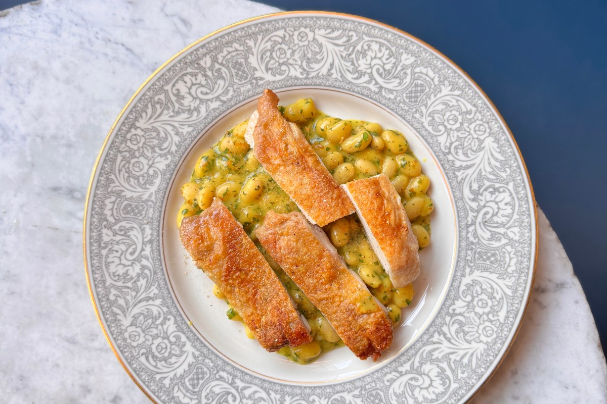 Overhead view of a plated dish: sliced seared chicken breast over white beans in herb sauce on a patterned plate.
