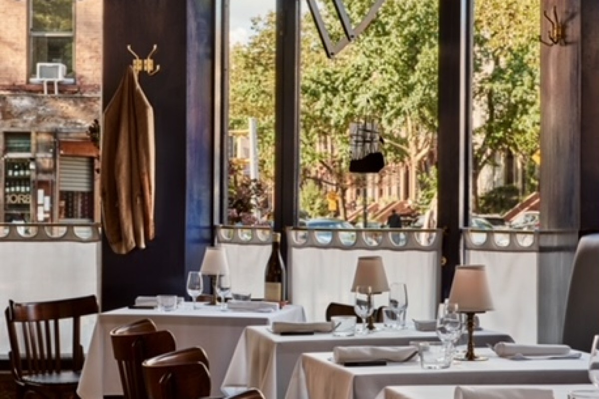 Restaurant dining room interior with white-clothed tables, lamps, and large windows overlooking a tree-lined street.