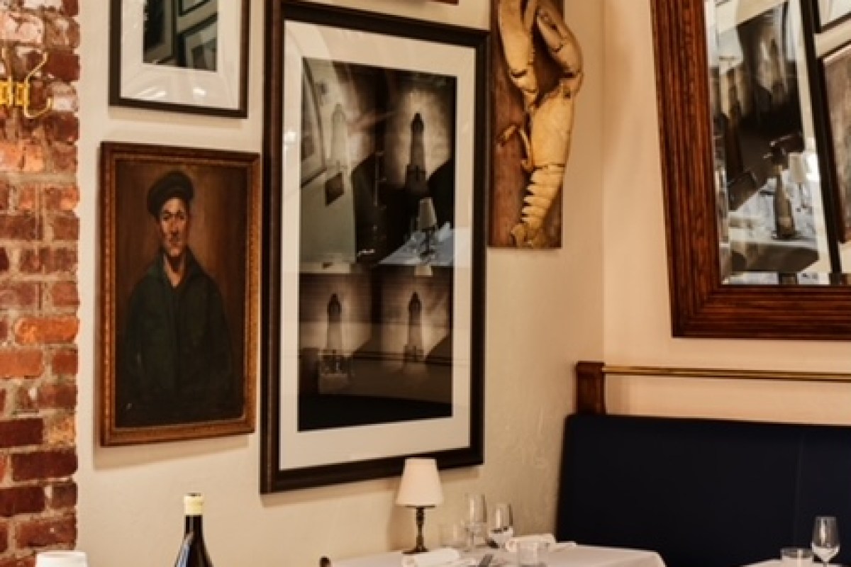 Dining room wall with framed artwork, a lobster sculpture, and a table set with wine glasses in the background.