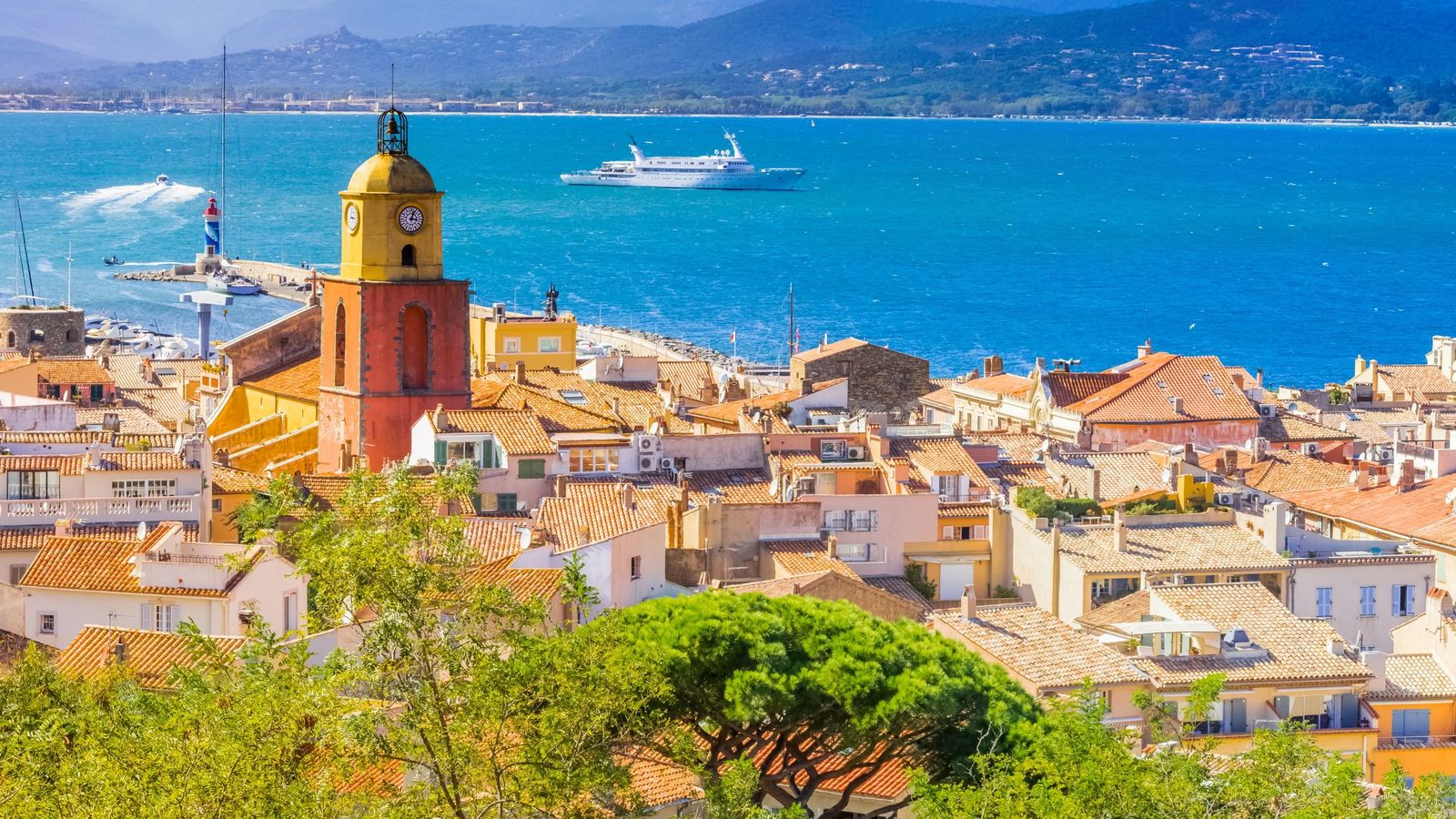 The yellow and red bell tower of Saint-Tropez above the rooftops, with the gulf and a yacht on the blue sea