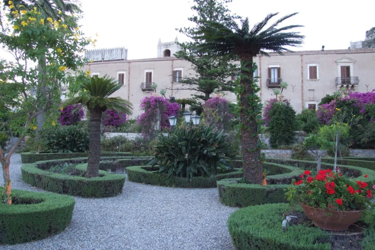 Manicured garden with trimmed hedges, palms and bougainvillea in front of the hotel's historic beige façade with shuttered windows.