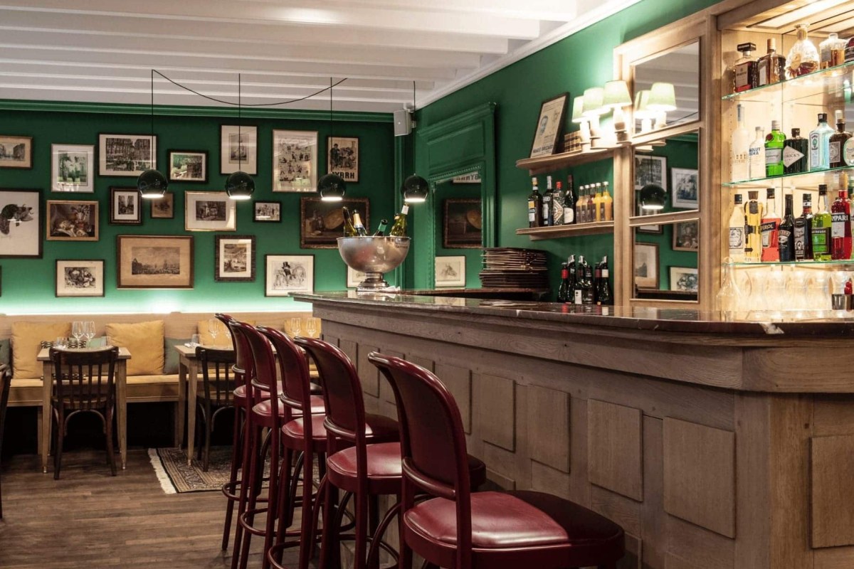 Restaurant bar with red stools, marble counter, bottles on shelves, and green gallery wall in background