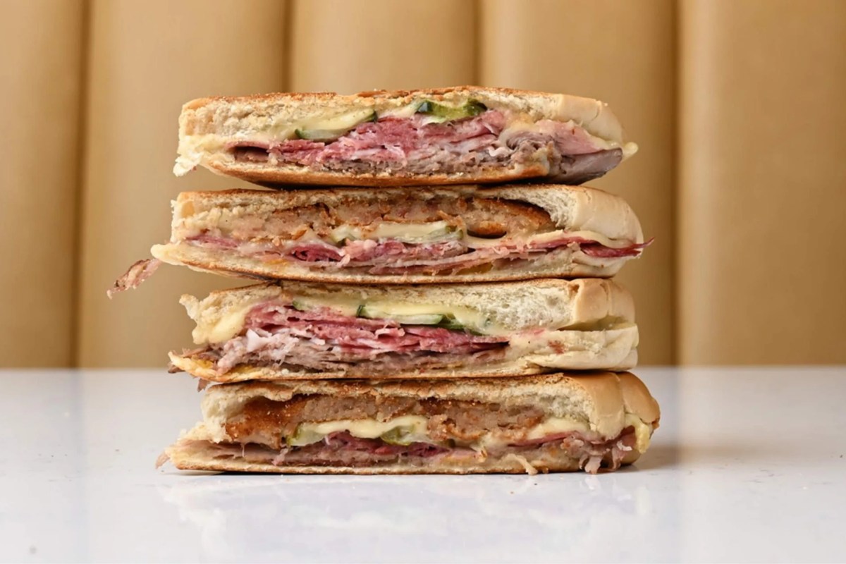 Stack of four halves of a pressed Cuban sandwich showing layered meats, cheese and pickles on a plain counter.