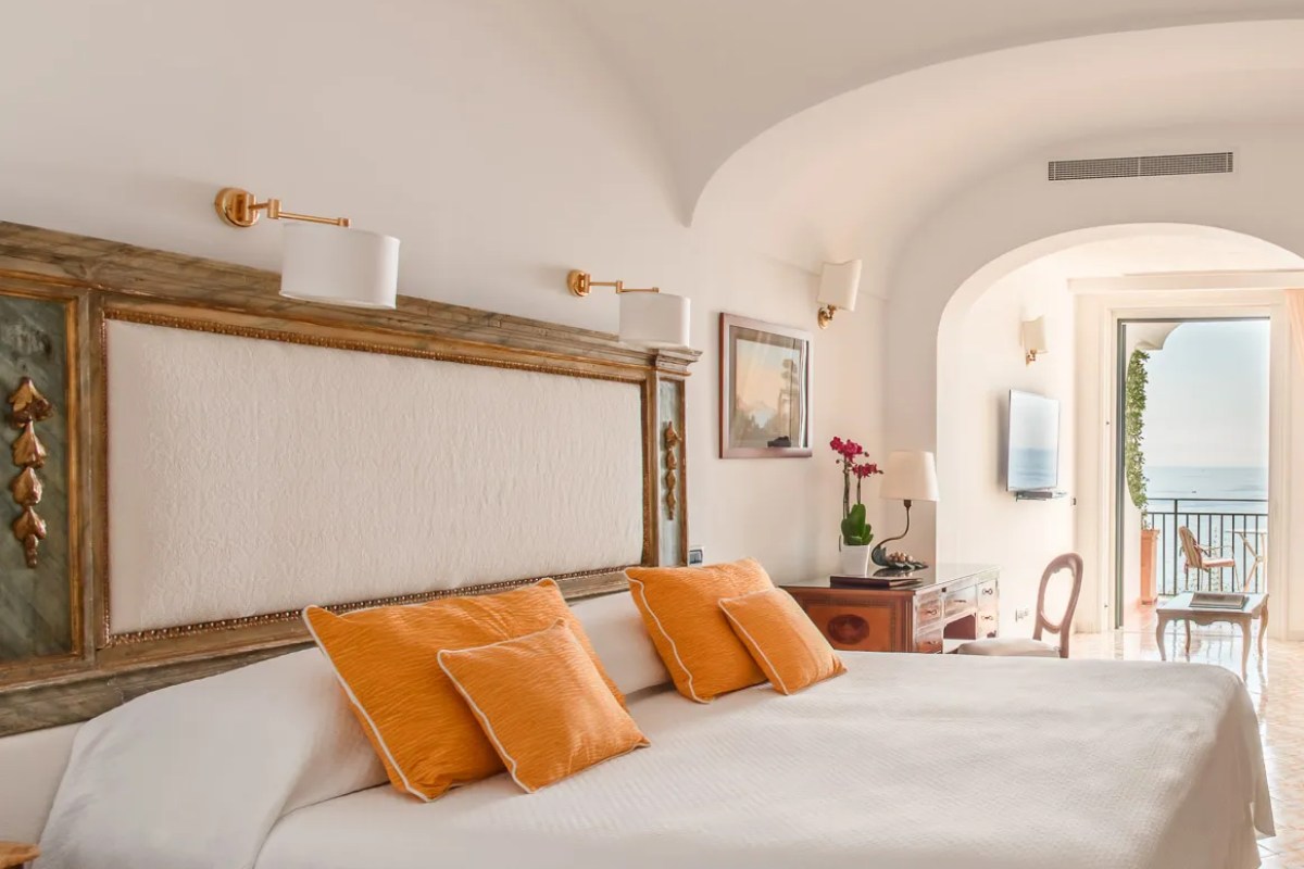 Hotel bedroom with orange pillows, gilded headboard, and balcony overlooking the sea