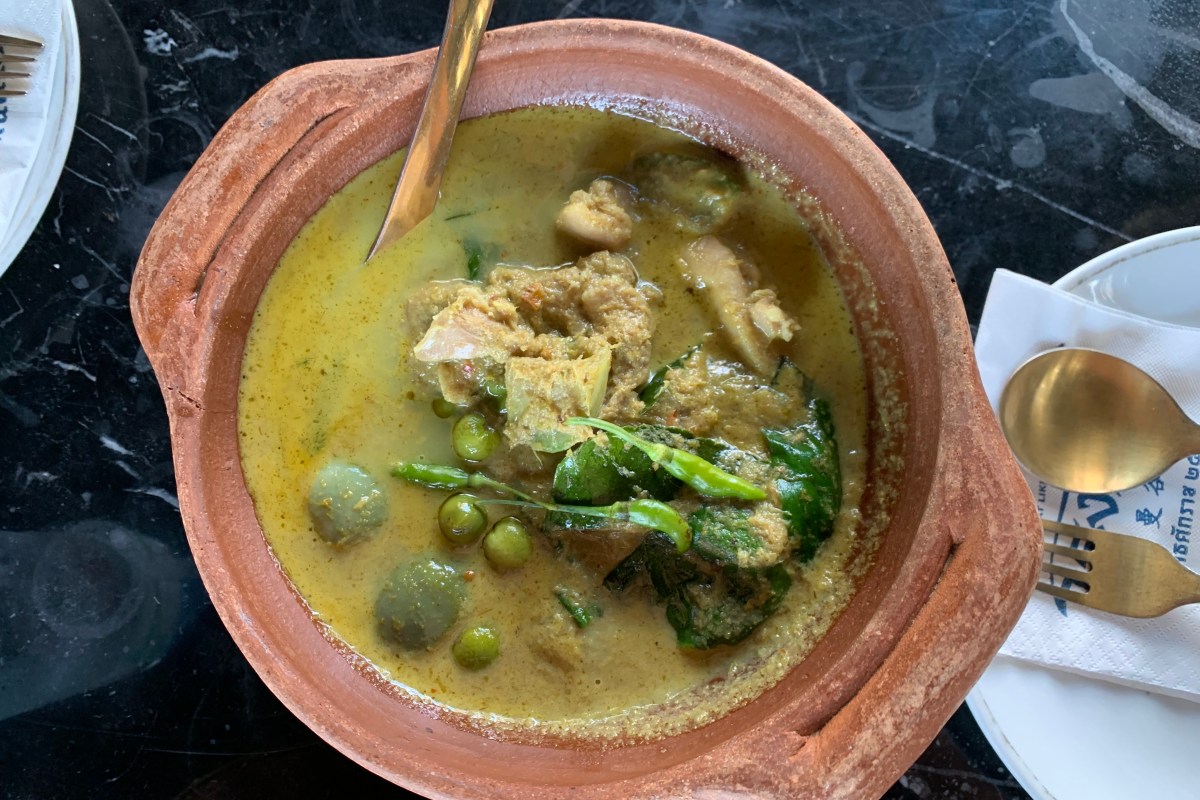 Overhead shot of a green curry with chicken, eggplant, and herbs in a clay bowl on a dark marble table
