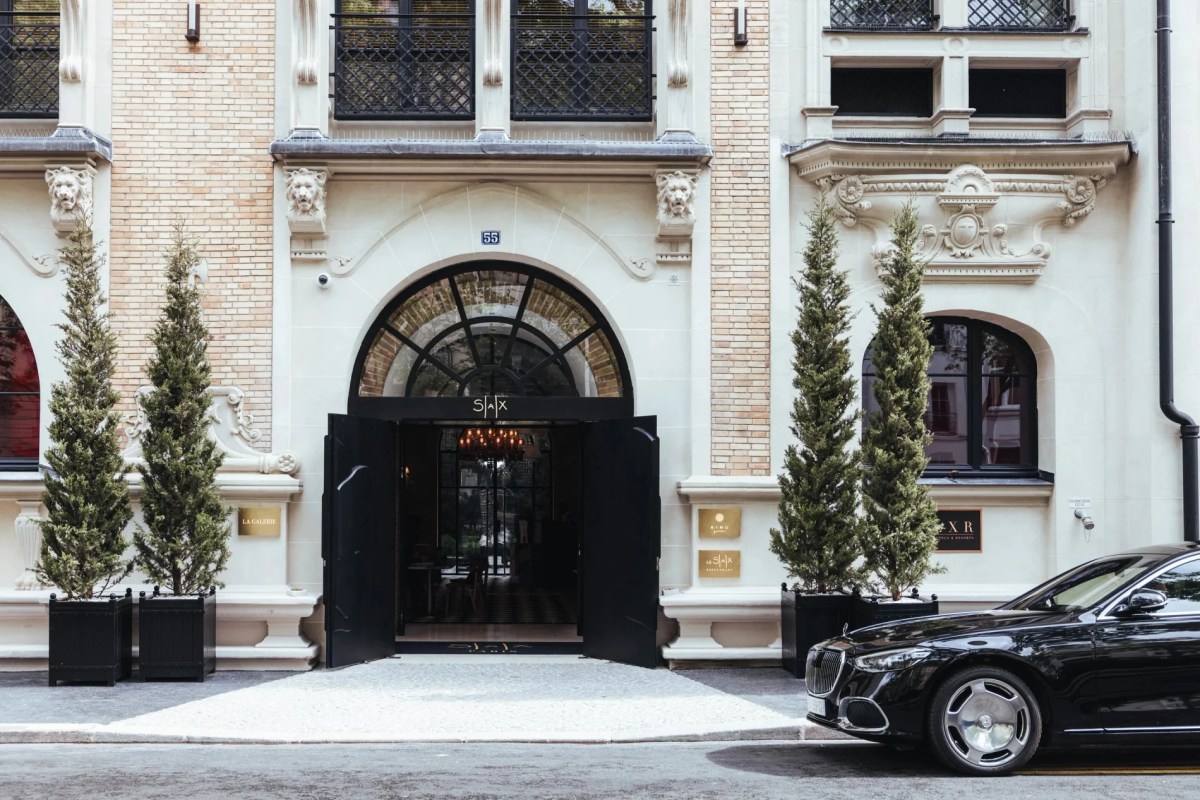 Hotel façade with ornate stone entrance, lion motifs, black doors, SAX signage, and topiary trees out front