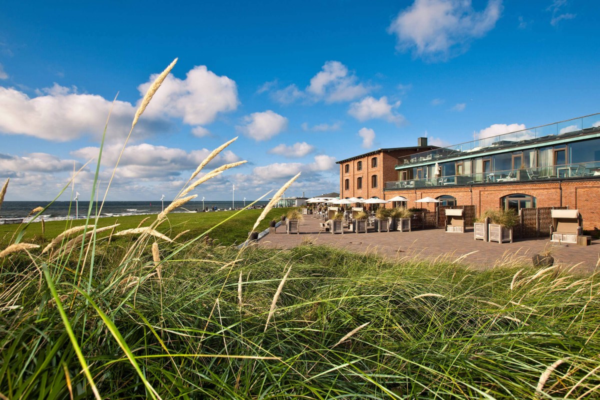 Daytime view of the red-brick Seesteg Norderney hotel with terrace, umbrellas and dune grass in foreground, sea in background.