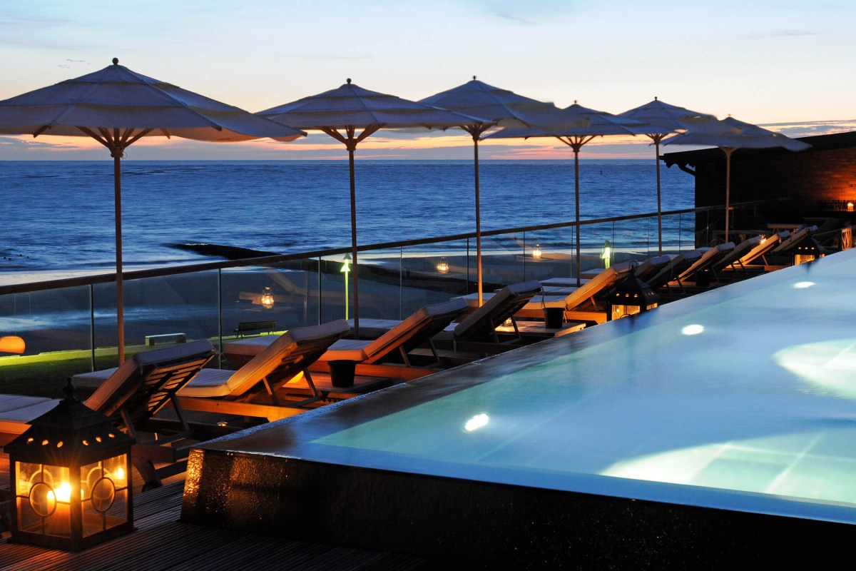 Rooftop infinity pool with loungers and umbrellas overlooking the sea at dusk, hotel building visible at right